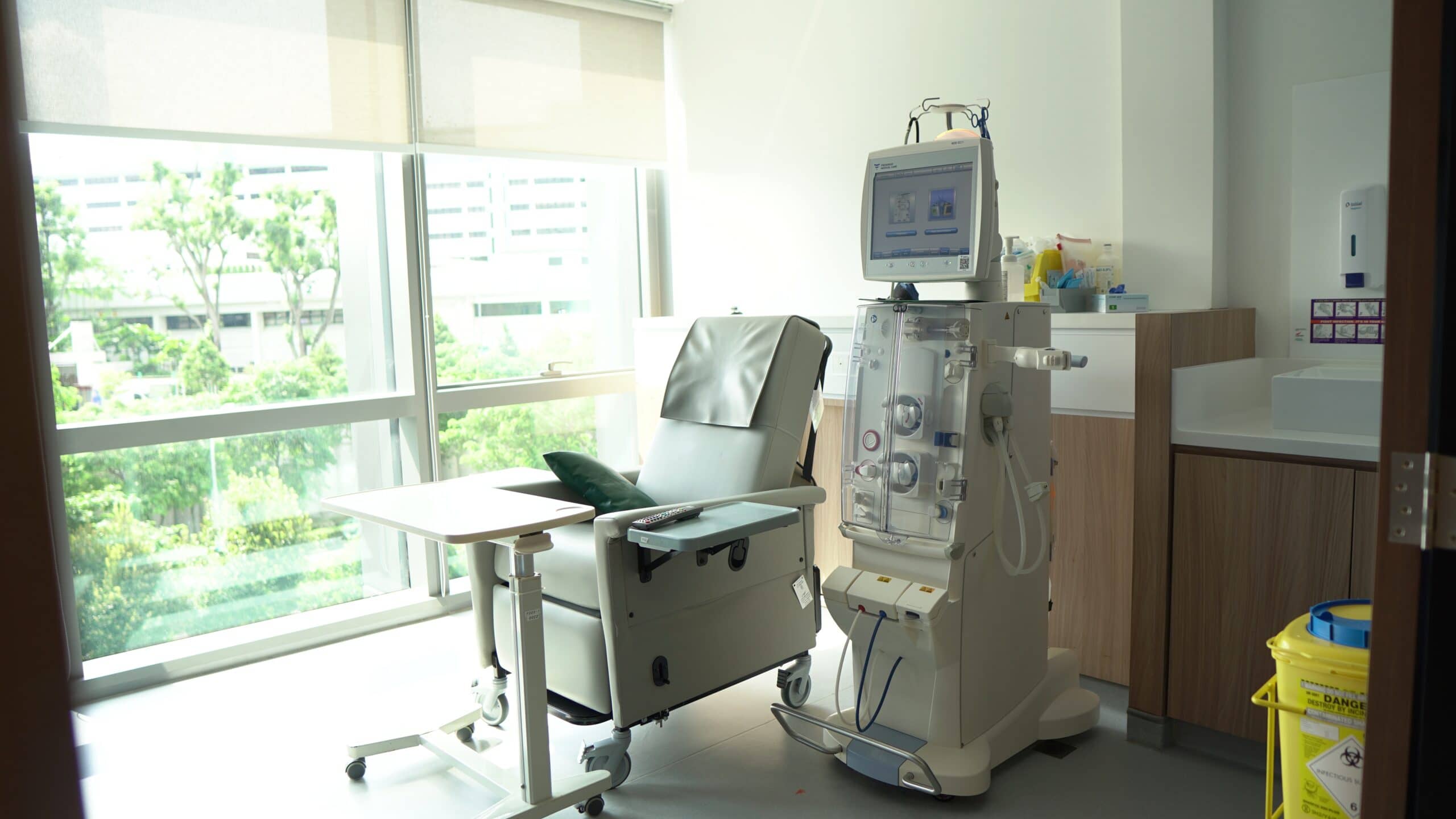 Bright dialysis room: chair, machine, bedside table by a large window.