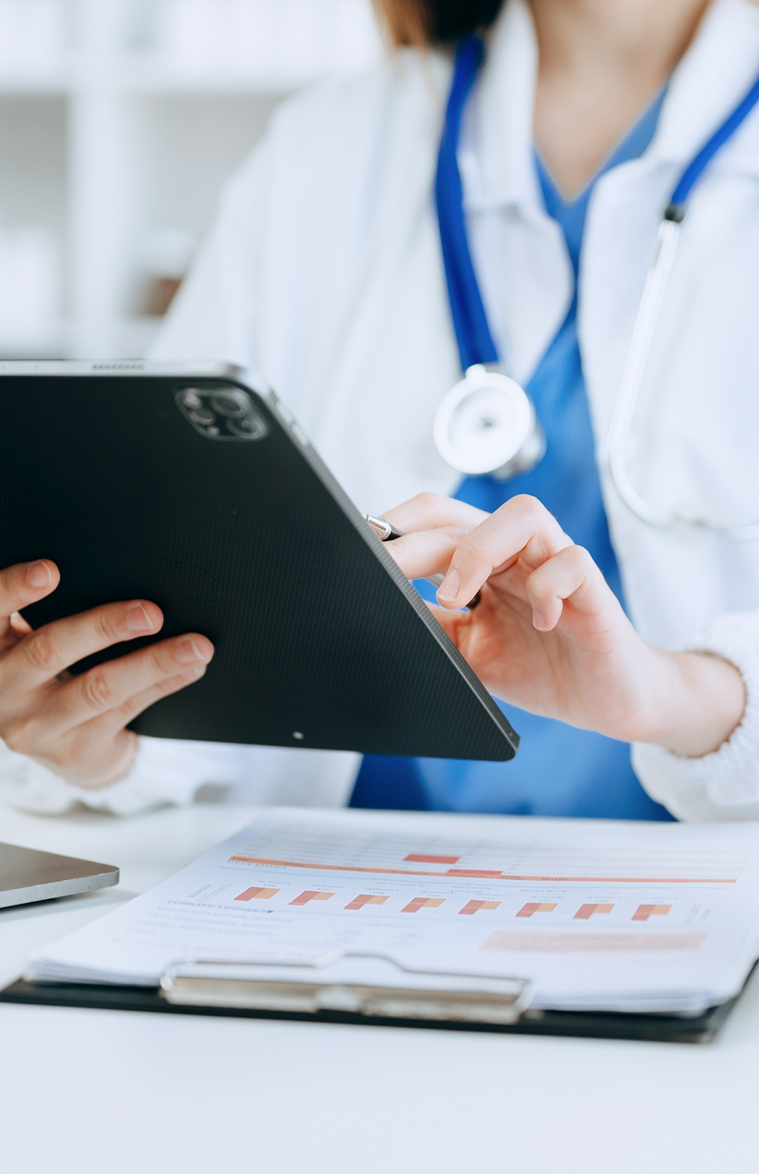 A medical professional in a lab coat and stethoscope uses a tablet, with charts on a clipboard in the foreground, indicating clinical data management.